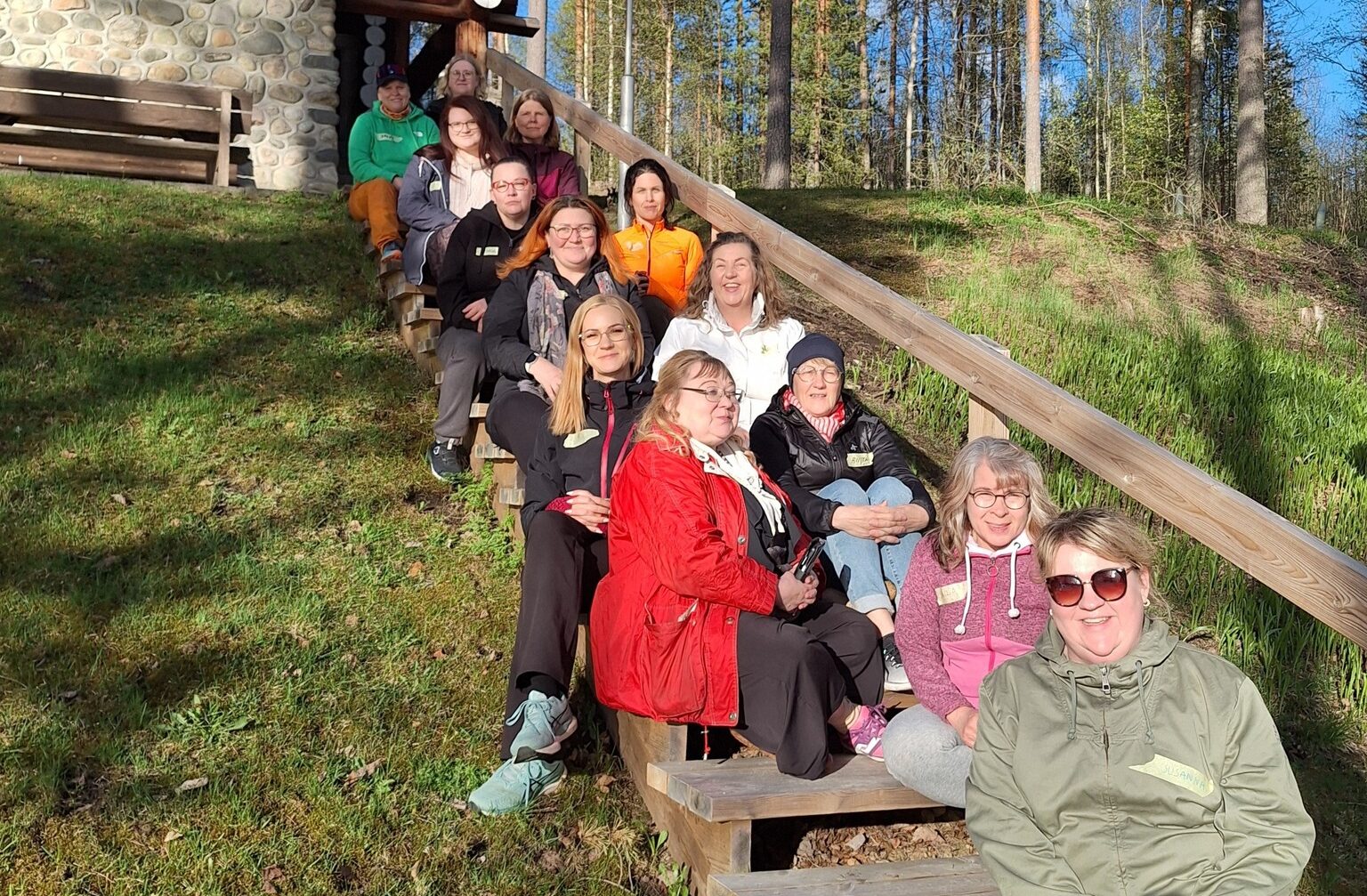 Yhdistyksen hallituksen jäseniä sekä jäseniä istuu metsäisessä maisemassa puisilla portailla auringon paisteessa.